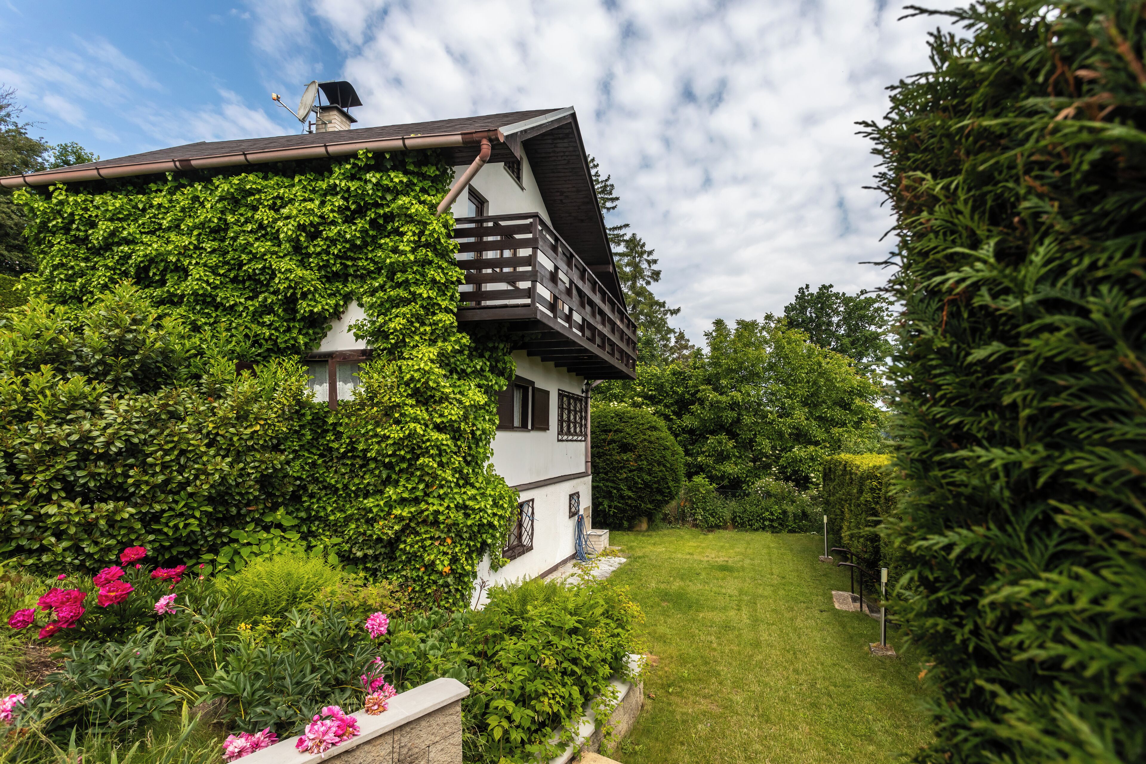 A house with a balcony, surrounded by lush greenery and a well-maintained lawn.