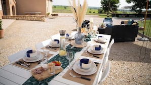 Un espace de restauration en plein air avec une table dressée pour deux personnes, agrémenté d’un mur en pierre rustique, d’une vue sur la campagne et d’un coin salon extérieur décontracté.