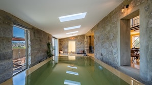 An indoor swimming pool with stone walls and skylights, a seating area with a view, and a dining area visible through an open doorway. - Lemon Villa-indoor Pool (Porto)