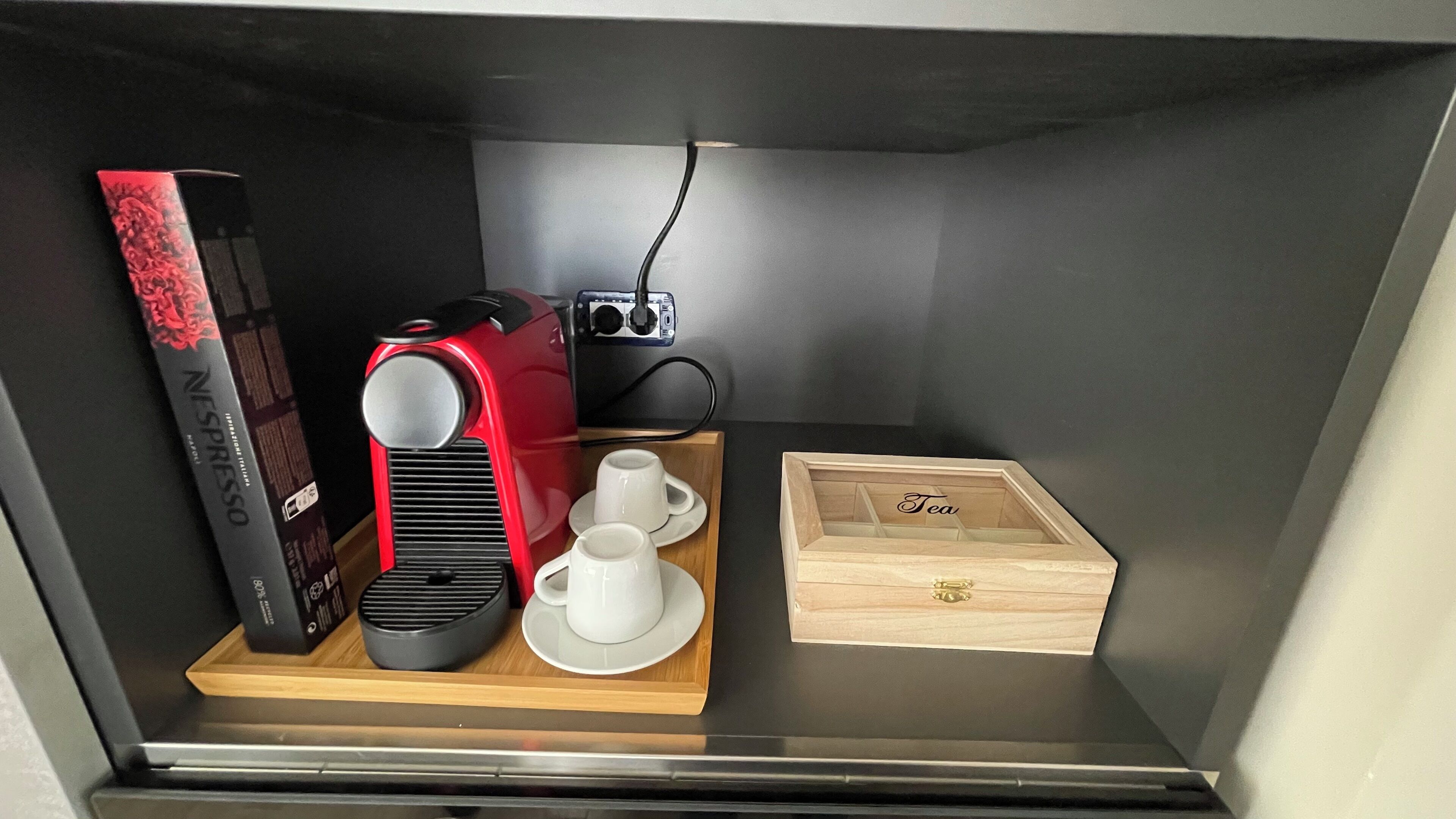 Un angolo cottura compatto con una macchina da caffè rossa, una cialda Nespresso, due tazze bianche, una piccola radio e una scatola di legno con l'etichetta 'Tè'.