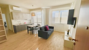 A modern apartment with a kitchen, dining area, and living room. It features a sofa, a dining table with chairs, a TV mounted on the wall, and a window with a view.