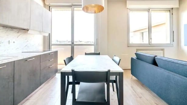 A modern kitchen with a dining table and chairs, a sofa, and large windows.