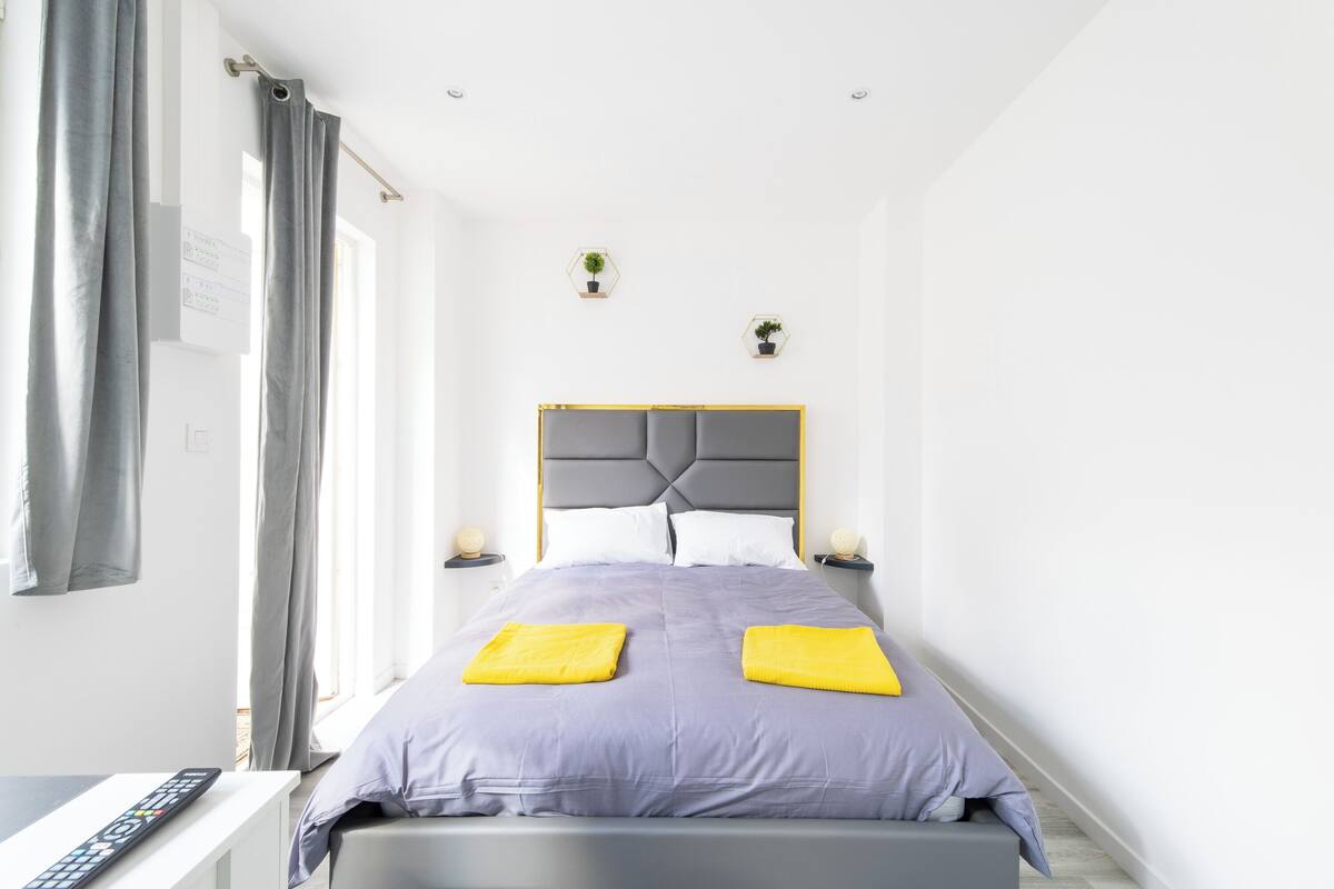 A modern bedroom with a grey headboard, two bedside tables with lamps, a bed with a purple bedspread and two yellow pillows, and a window with grey curtains.