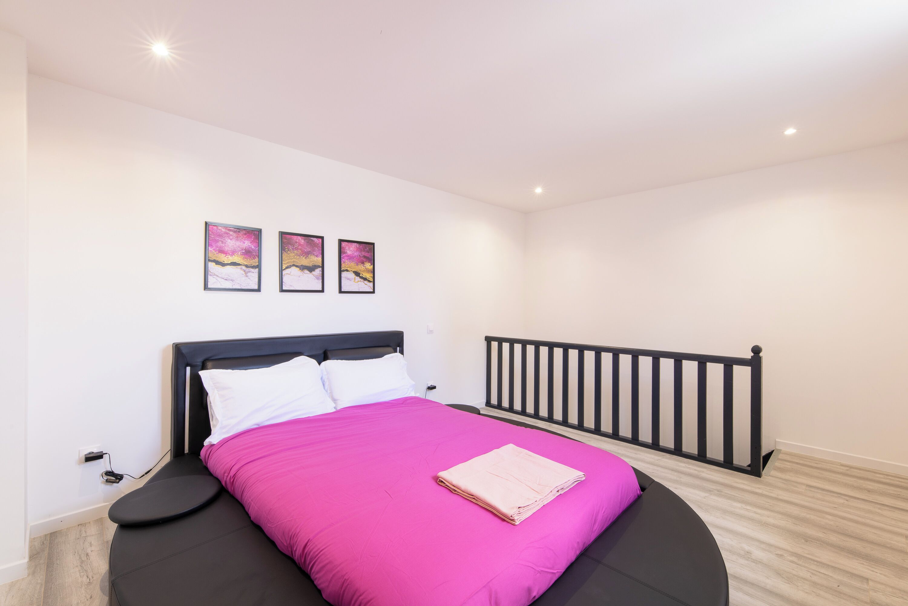 A bedroom with a bed, a black headboard, a white wall, and a wooden floor. There are three framed pictures on the wall.