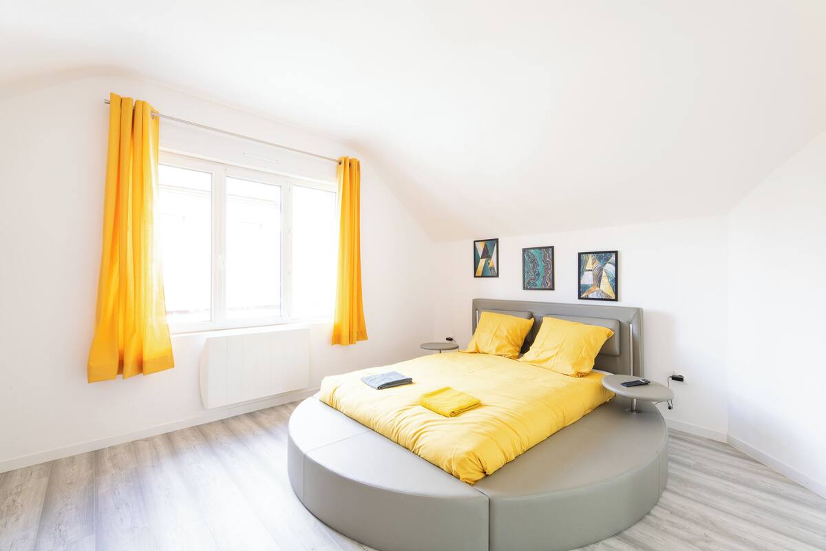 A modern bedroom with a curved bed, a bedside table, and a window with yellow curtains.
