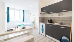 A modern kitchen with dark cabinets, a sink, and a countertop. There is a dining area with a table and chairs, and a bed with a blue and white pattern in the background. -  GLM Residences 1 (Saint-Quentin)