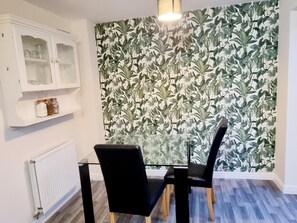 A dining area with a glass table, black chairs, a white cabinet with glass doors, and a wall with a leafy wallpaper pattern.