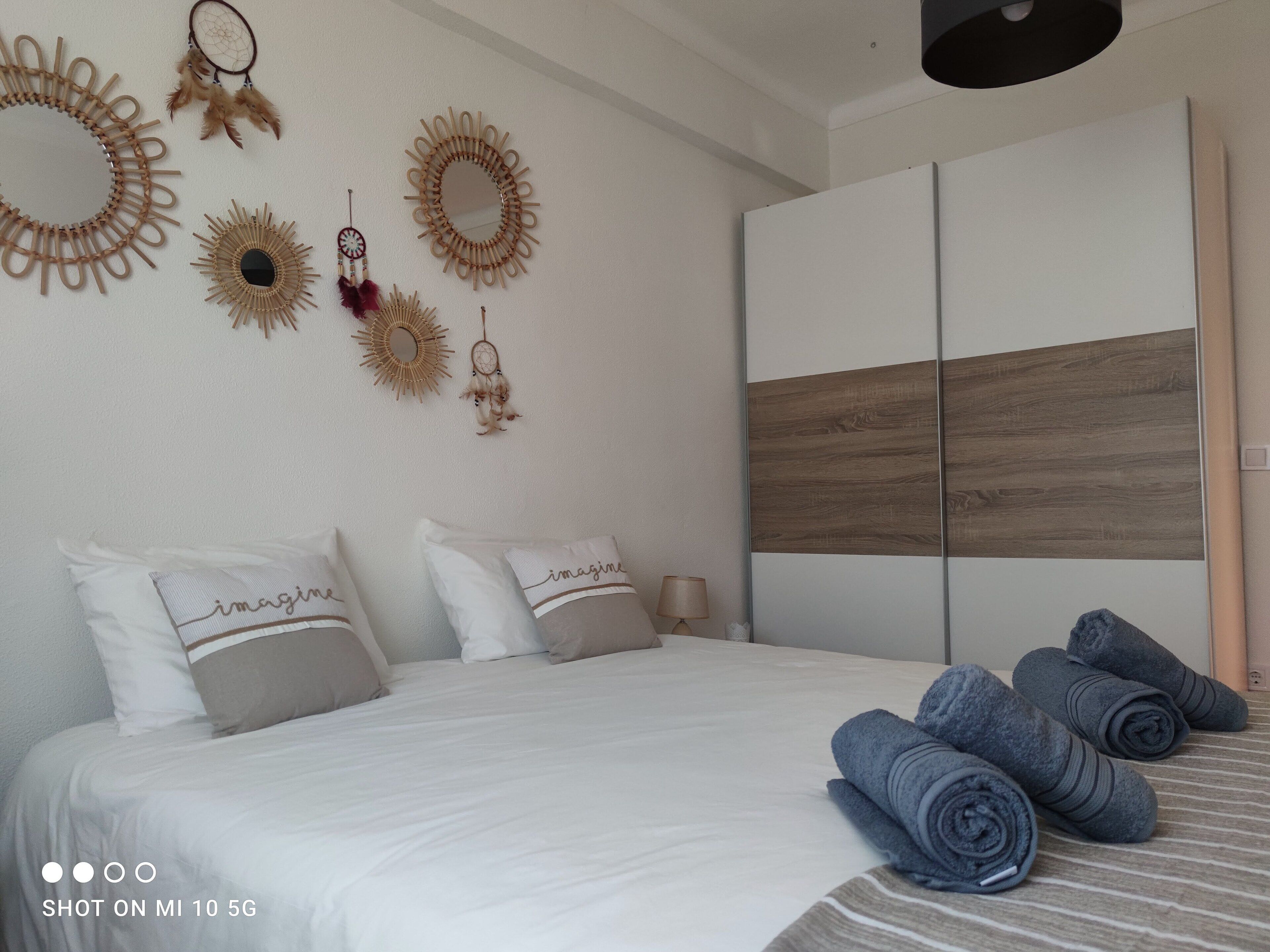Ein ordentlich bezogenes Bett mit weißen und beigen Kissen, einem hölzernen Kopfteil und einem großen Spiegel. Auf dem Bett befinden sich Wanddekorationen und aufgerollte Handtücher.