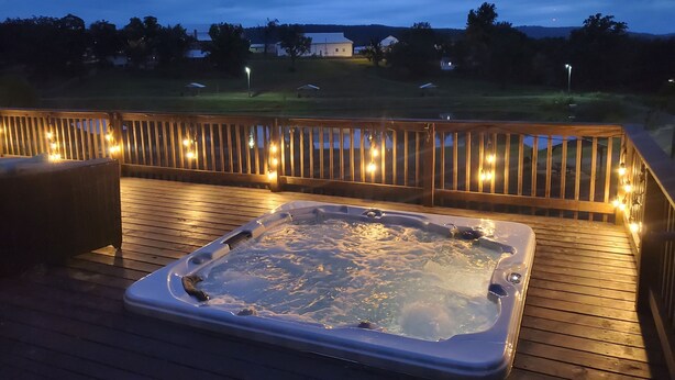 A hot tub on a wooden deck with string lights, surrounded by a wooden railing. - William's Catch at Racoon Springs - 16+ Guests (Marshall)