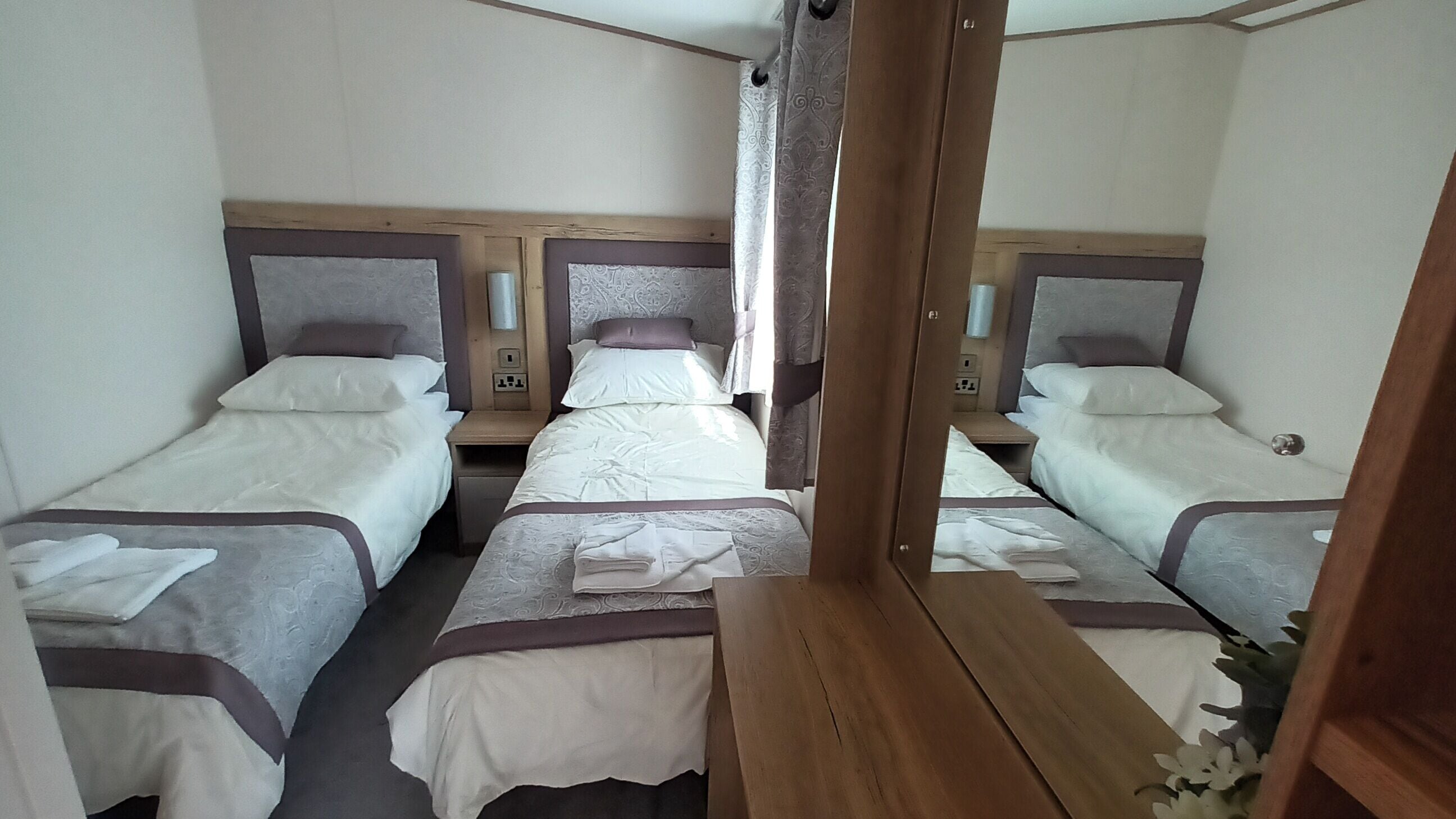 A hotel room with three single beds, wooden headboards, bedside tables, and a mirror.