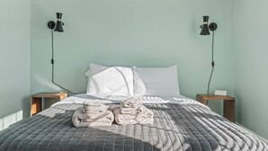 Un lit bien fait, recouvert d’une couette grise, avec deux oreillers blancs et des serviettes pliées, le tout adossé à un mur vert clair.