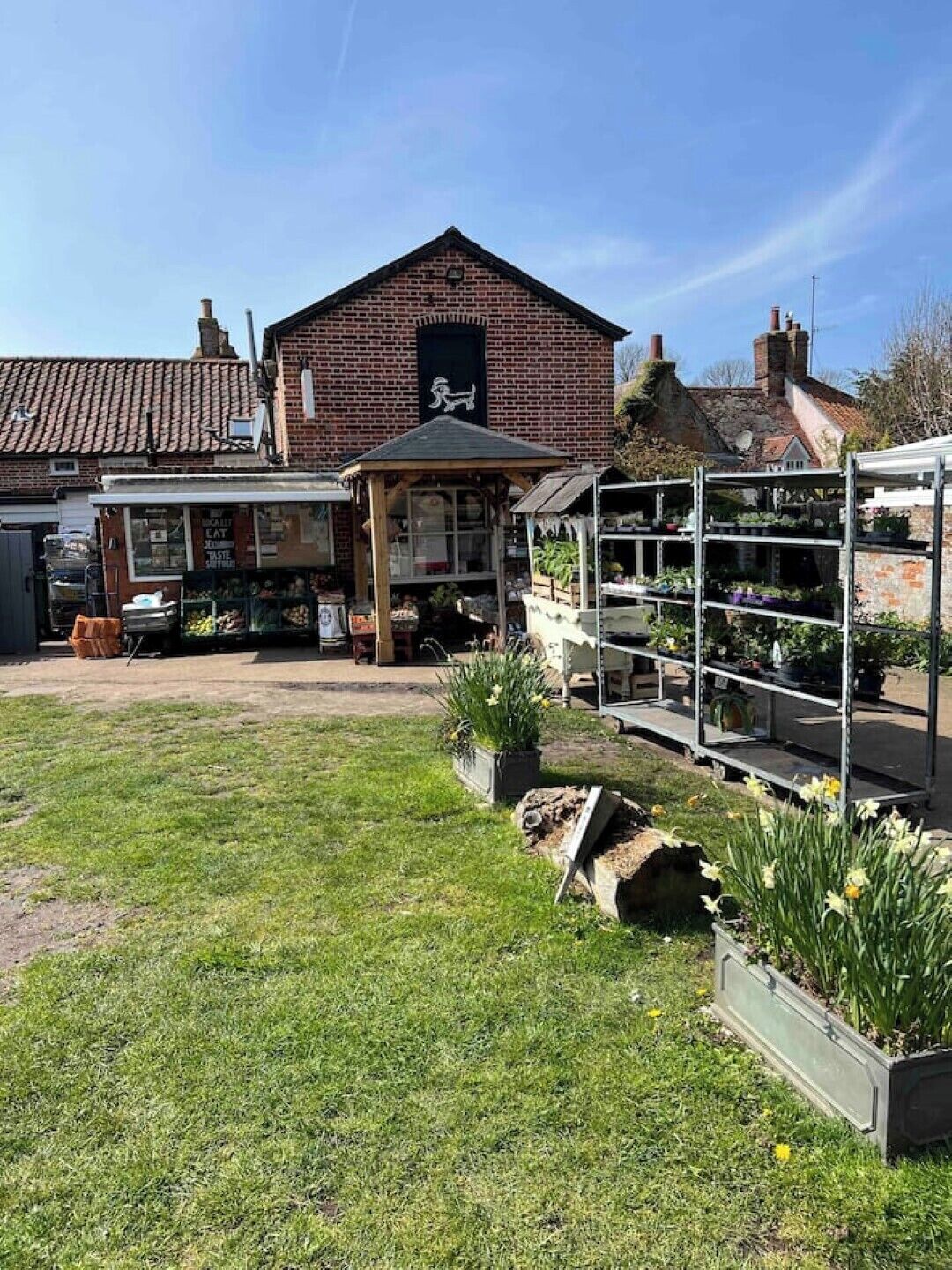 A brick building with a signboard, a covered outdoor seating area, and a garden with potted plants and flowers.