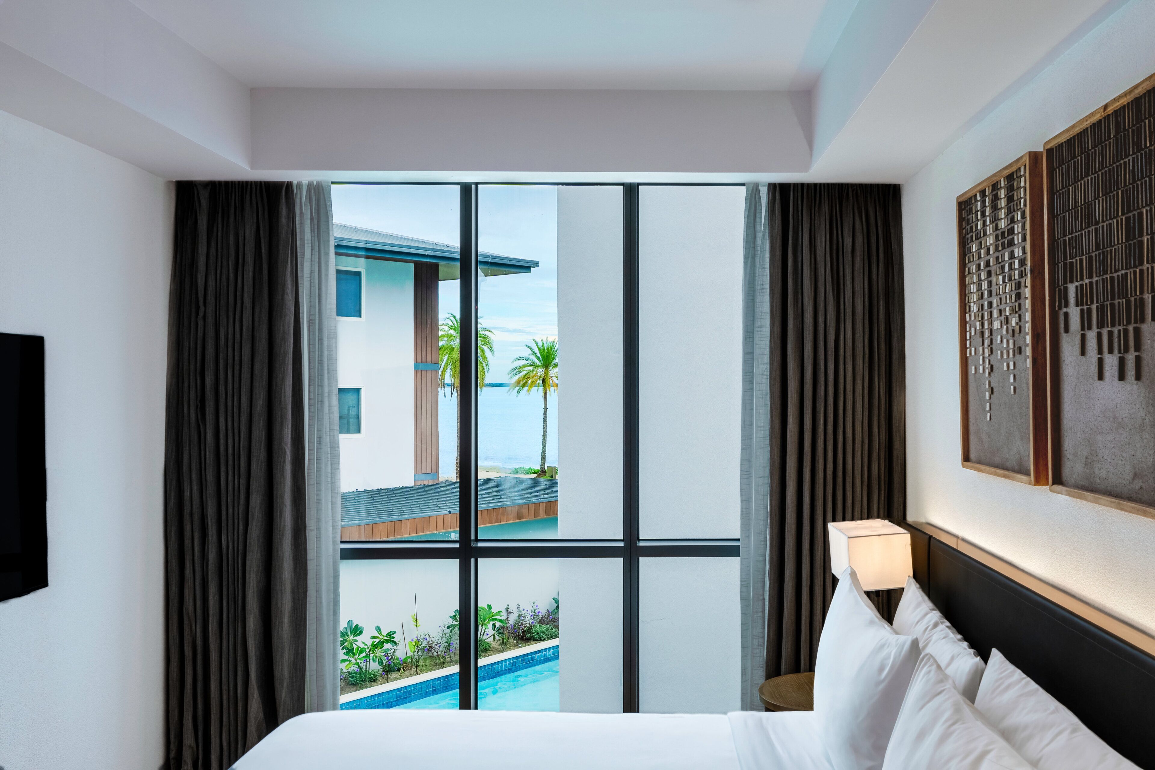 A hotel room with a bed, a TV, a window with curtains, and a view of a building and pool.