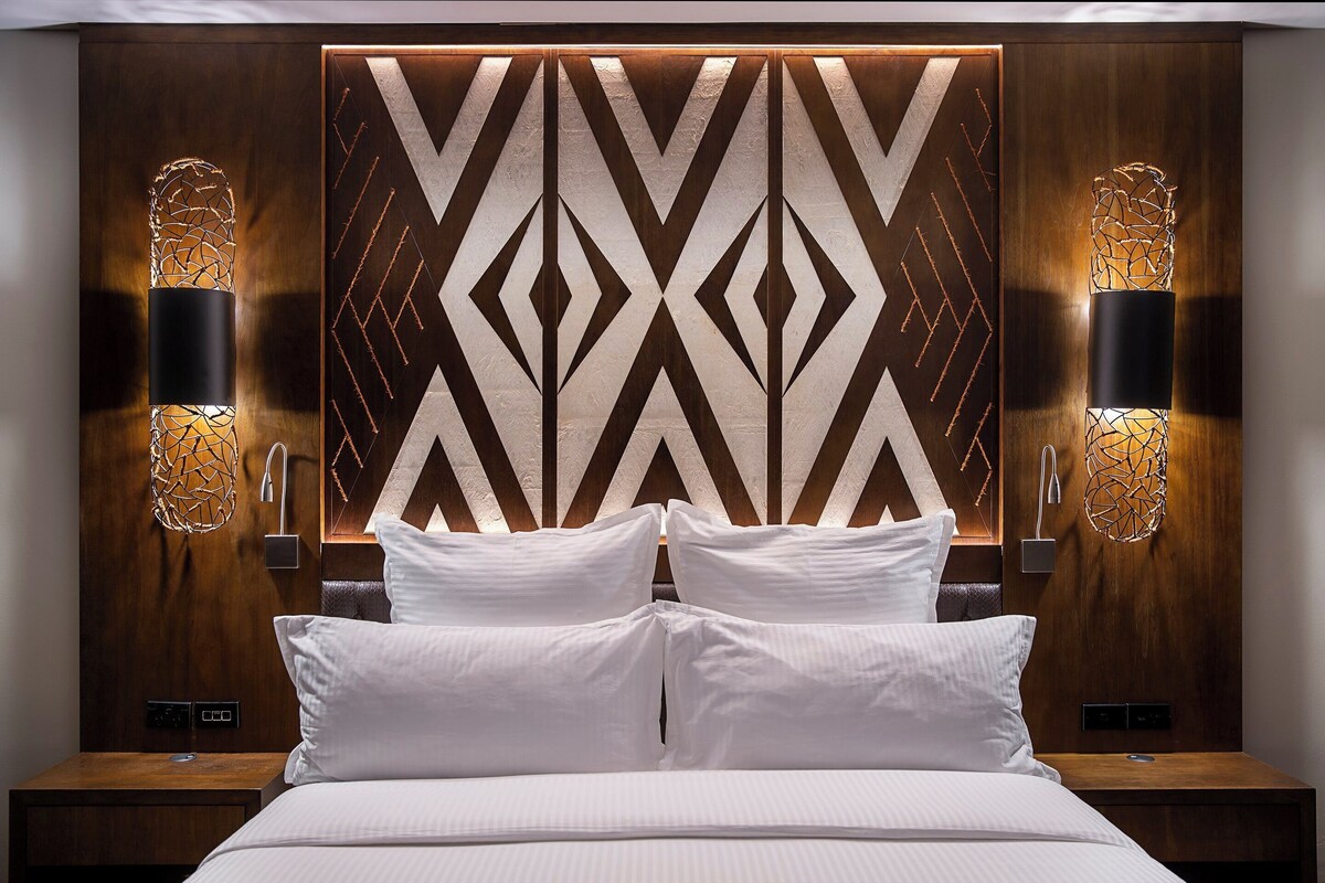 Una habitación de hotel moderna con una cama grande, dos mesitas de noche y un cabecero con diseño geométrico.