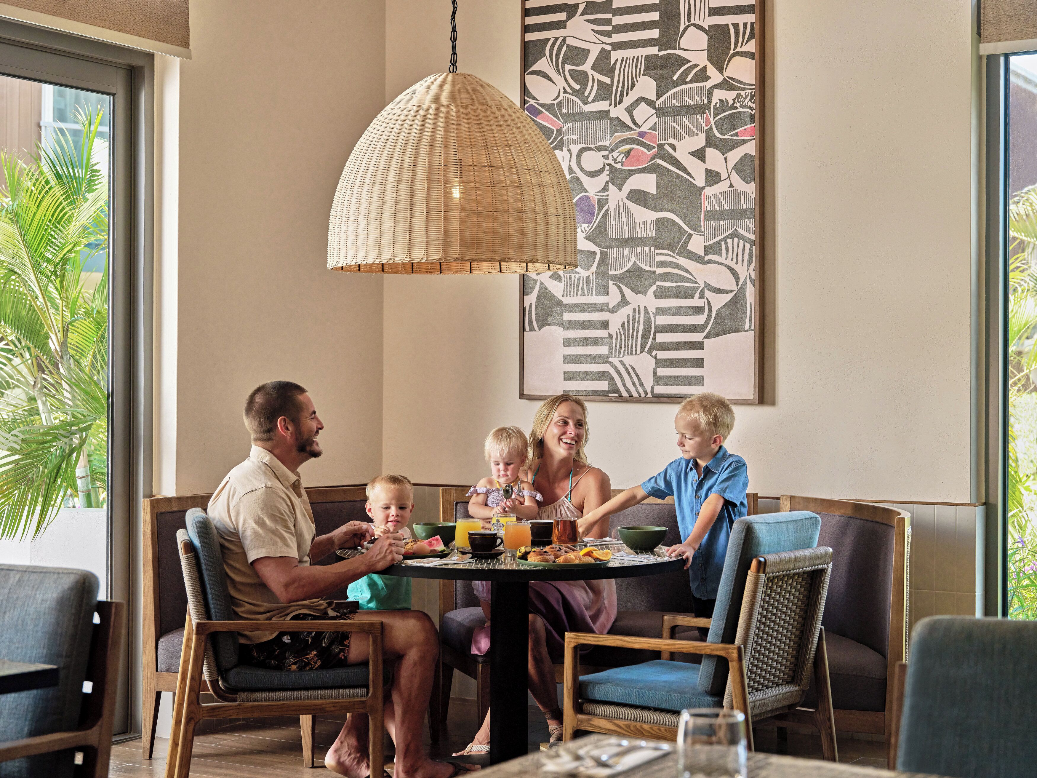 A family dining at a table with a woven pendant light, a large abstract painting, and a view of greenery outside.