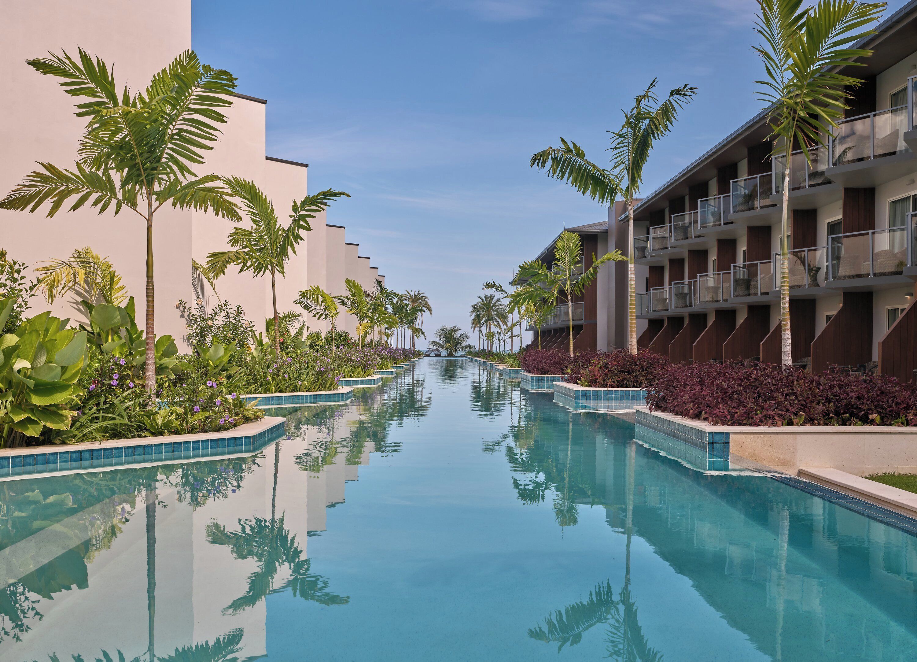A pool with clear water, surrounded by palm trees and plants, with modern buildings on either side.