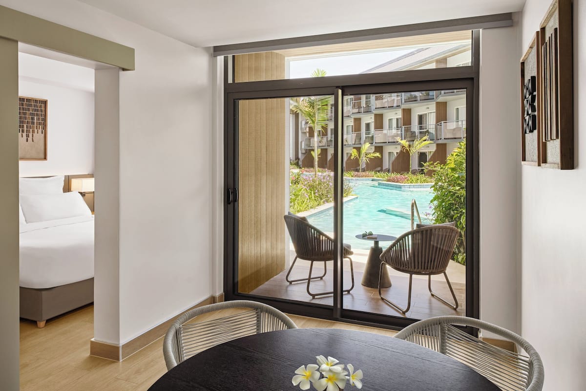 A hotel room with a sliding glass door leading to a pool area, a bed, a table with chairs, and a vase with flowers.