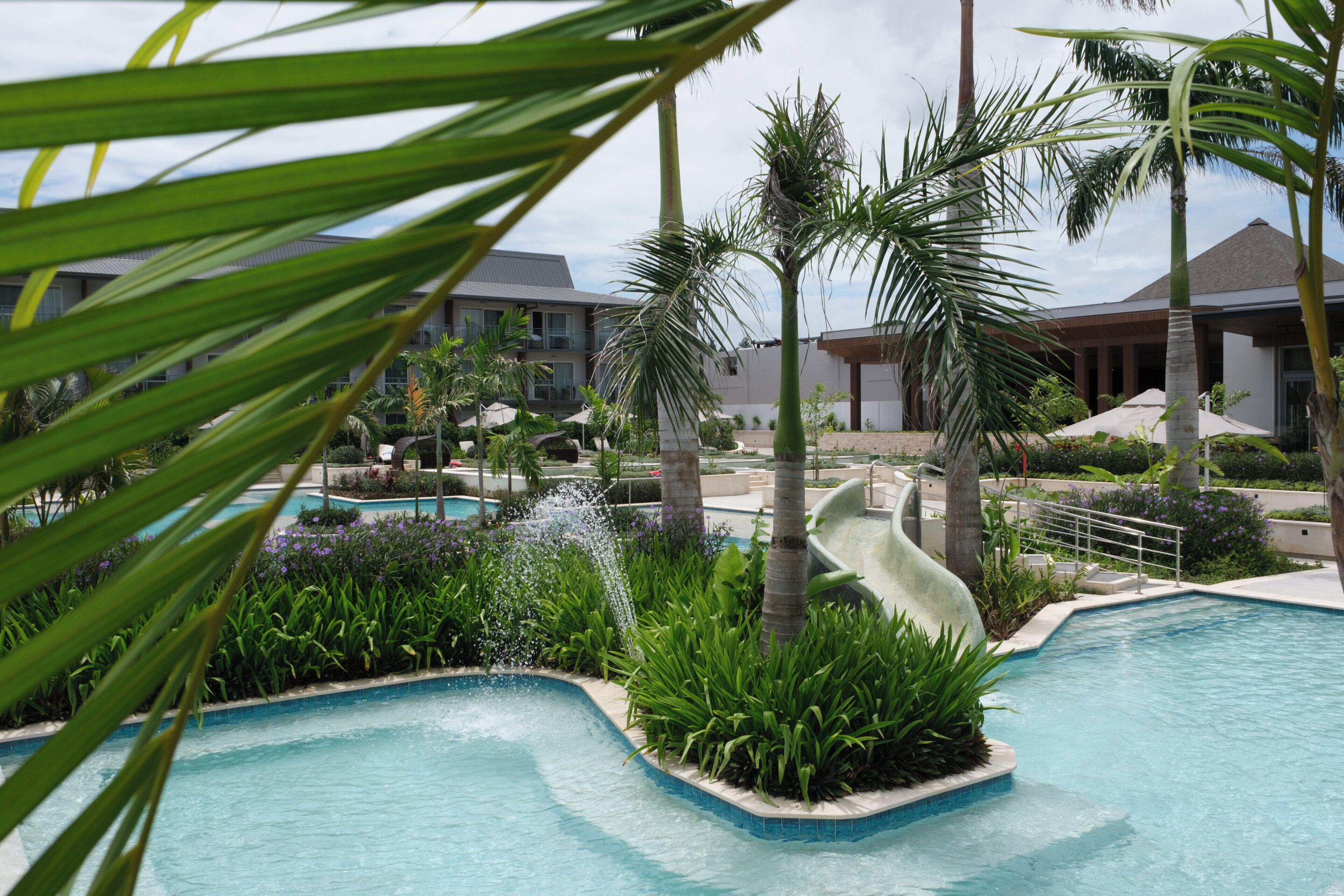 Área de piscina en un resort con tobogán de agua, palmeras y un edificio con balcones.