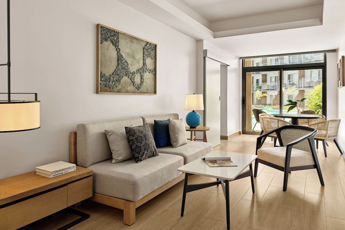 A modern living room with a sofa, coffee table, and chairs, a painting on the wall, and a view of the outside through large windows.