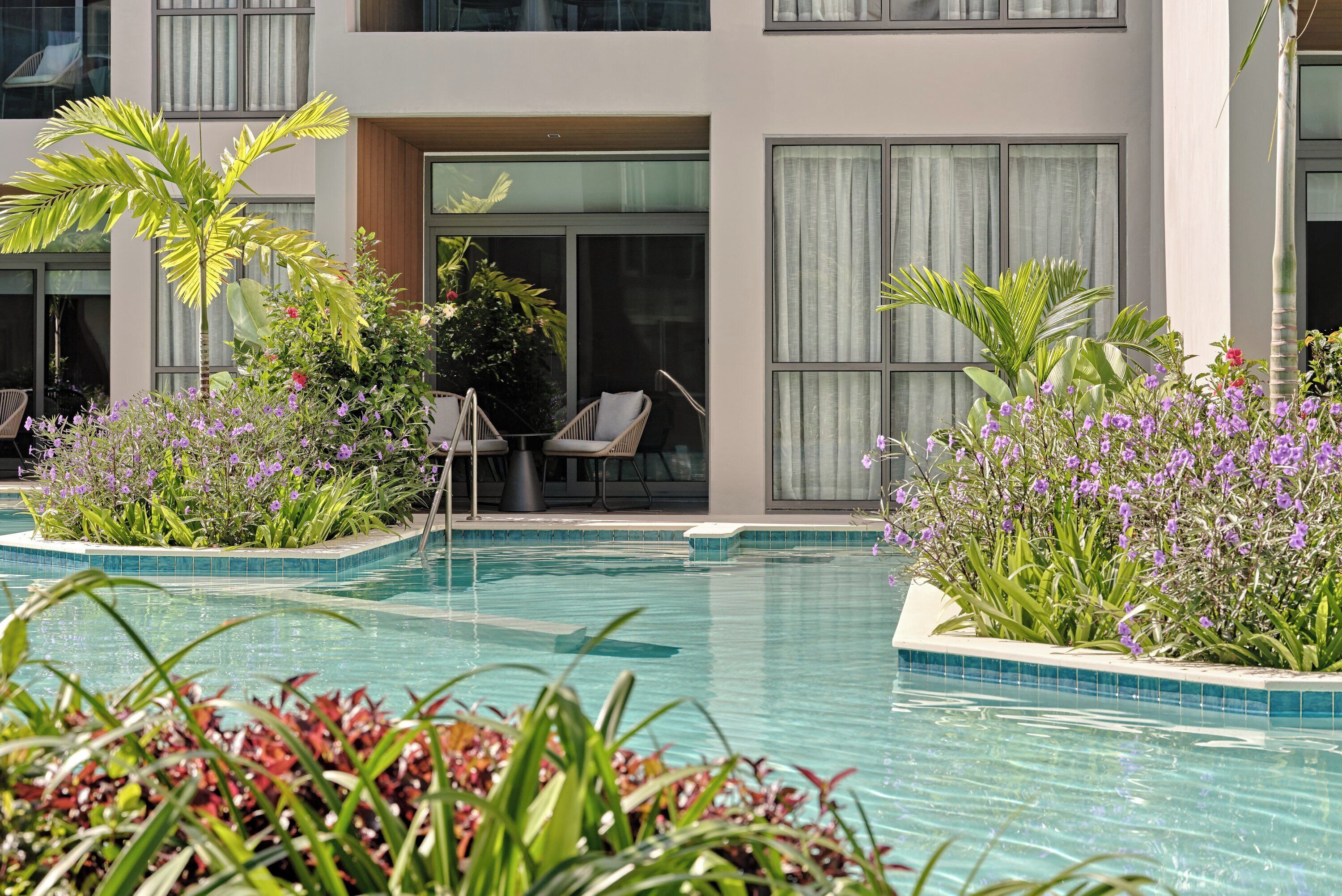 A modern hotel with a swimming pool surrounded by lush greenery and purple flowers.