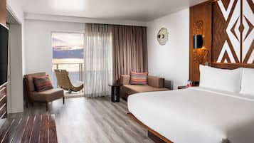 A hotel room with a large bed, a sofa, a chair, a small table, and a view of the sea through the window.