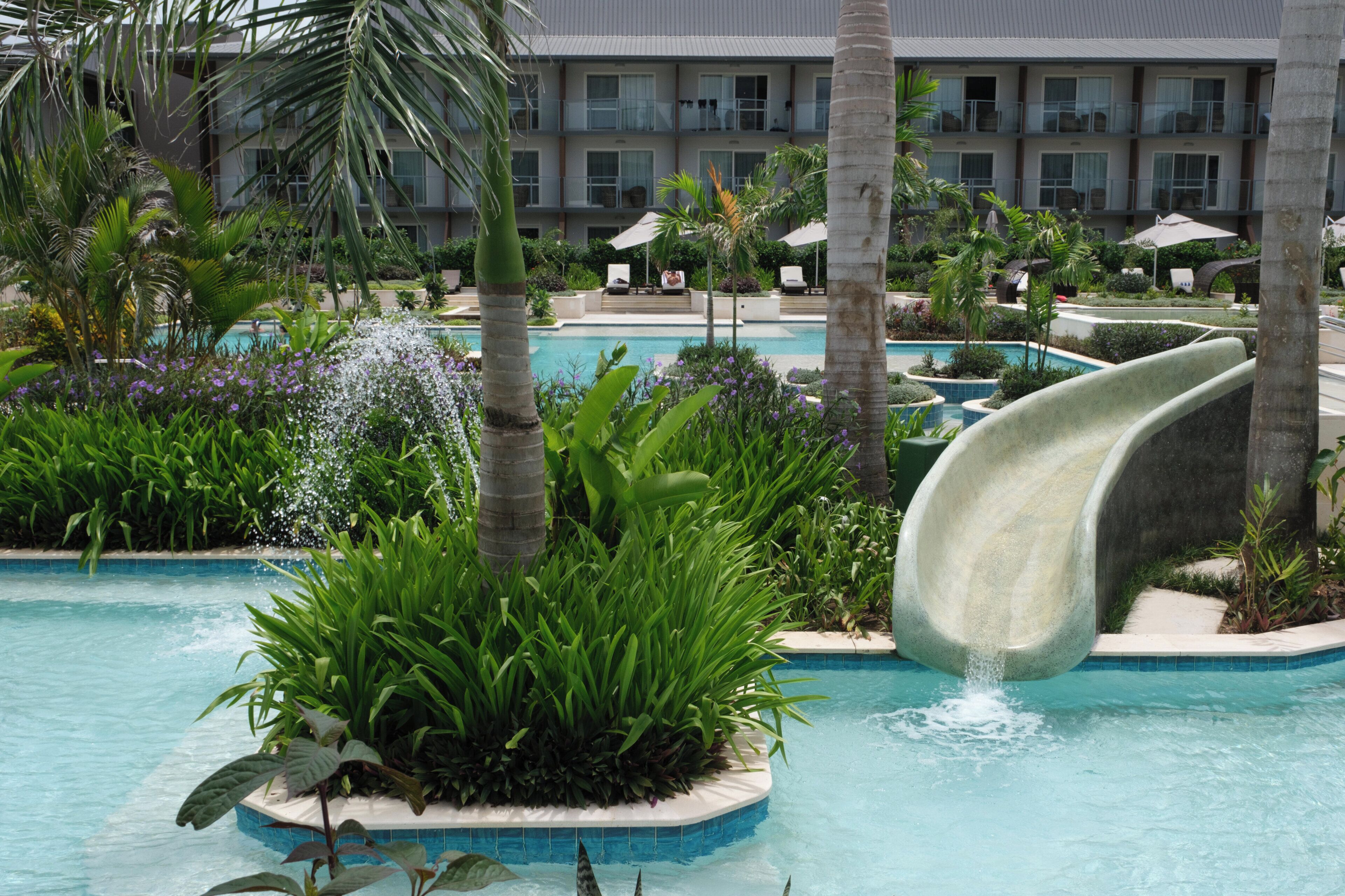 Un área de piscina con tobogán acuático, rodeada de abundante vegetación y palmeras.