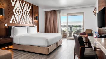 A hotel room with a large bed, a desk, a chair, a TV, and a balcony with a view.