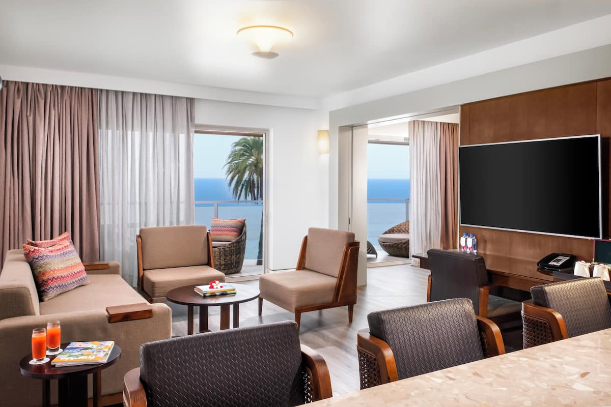 A hotel room with a sofa, armchairs, a small table, a TV, and a balcony with a view of the ocean.