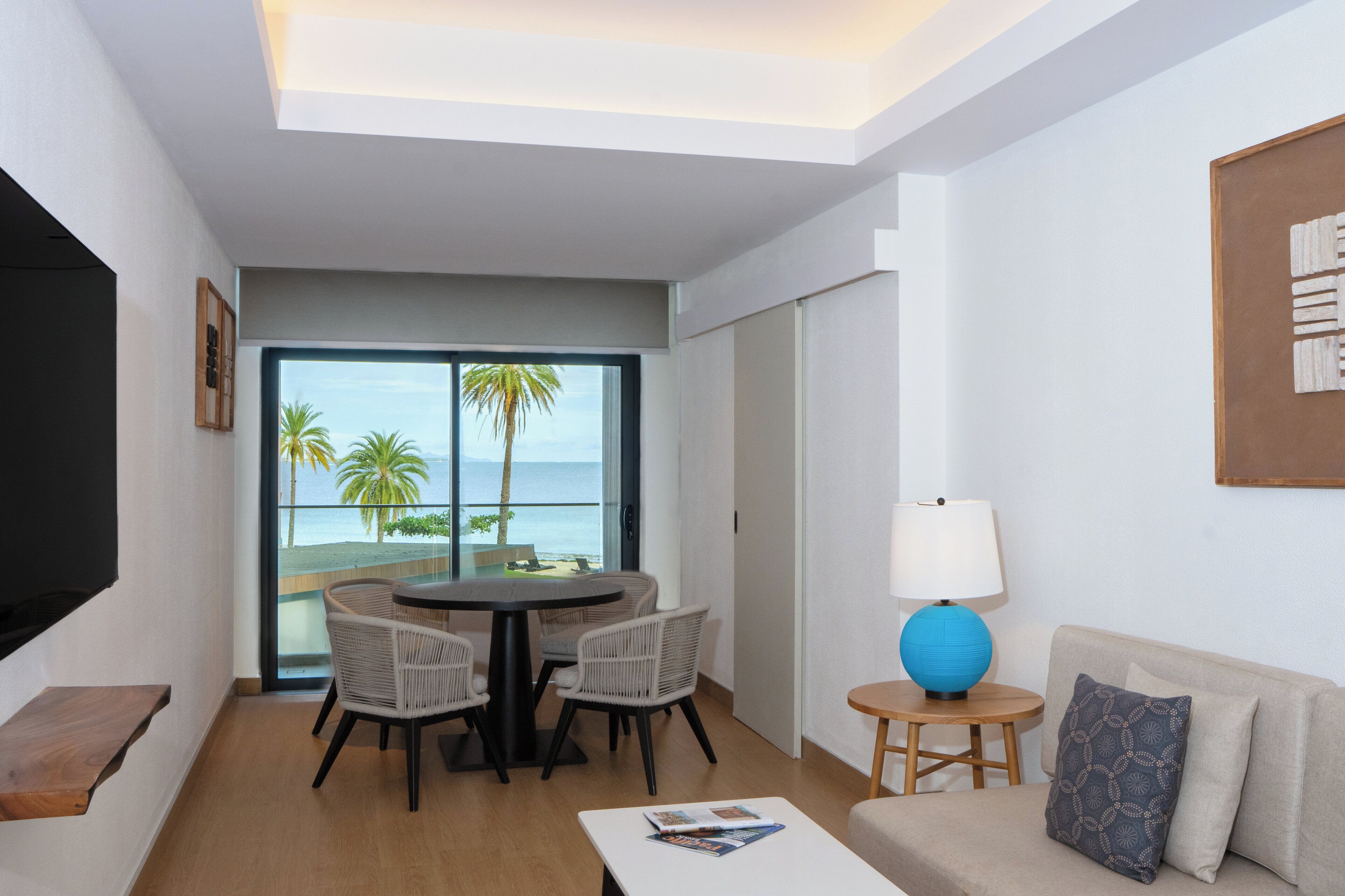 A hotel room with a balcony, a dining table with chairs, a sofa, a lamp, and a flat-screen TV.