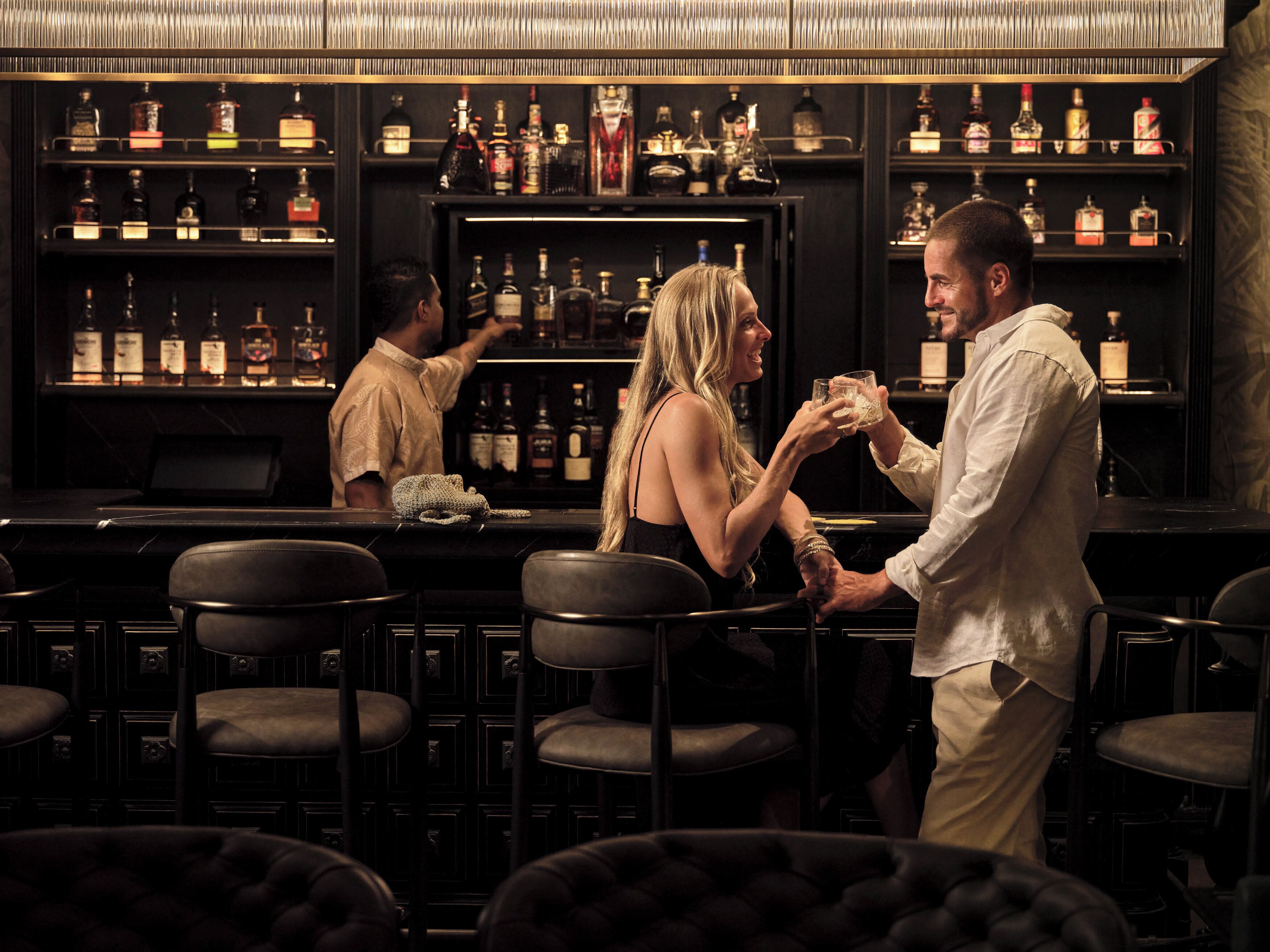 Uma área de bar com várias prateleiras de garrafas de bebidas, banquetas e um bartender.