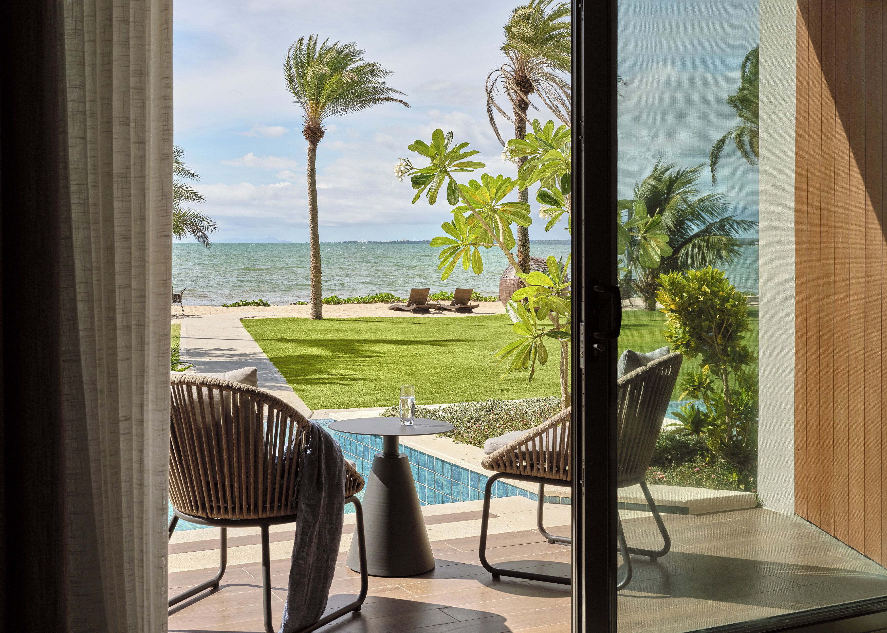 Blick aus einem Zimmer mit Glastür auf einen Poolbereich, mit Pool, Stühlen und einer Rasenfläche mit Palmen.