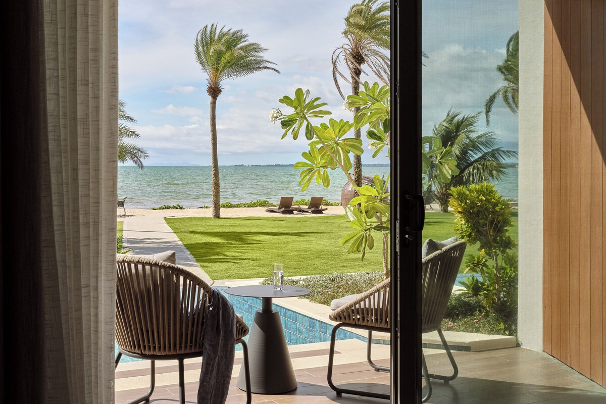 Vista desde una habitación con puerta de cristal a zona de piscina, que incluye piscina, sillas y césped con palmeras.