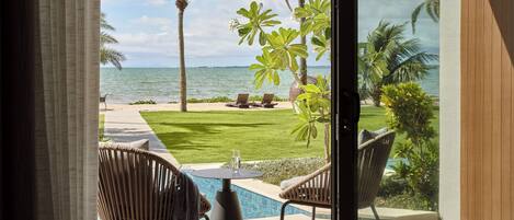 A view from a room with a glass door to a pool area, featuring a pool, chairs, and a lawn with palm trees.