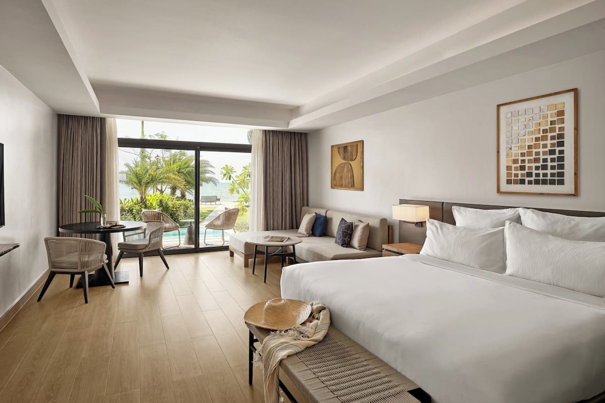 A hotel room with a large bed, a sofa, a dining table with chairs, and a view of the outdoors.