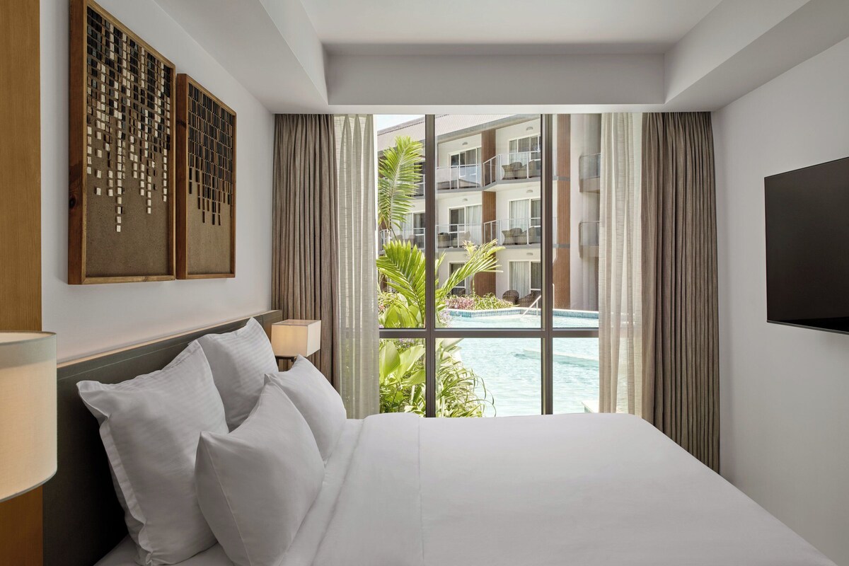 Habitación de hotel con una cama grande, televisor montado en la pared y vistas a una piscina y un edificio.