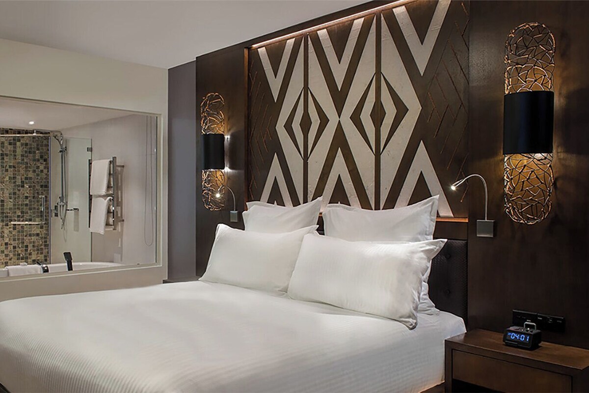 Una habitación de hotel moderna con una cama grande, un baño con ducha de vidrio y apliques decorativos en la pared.