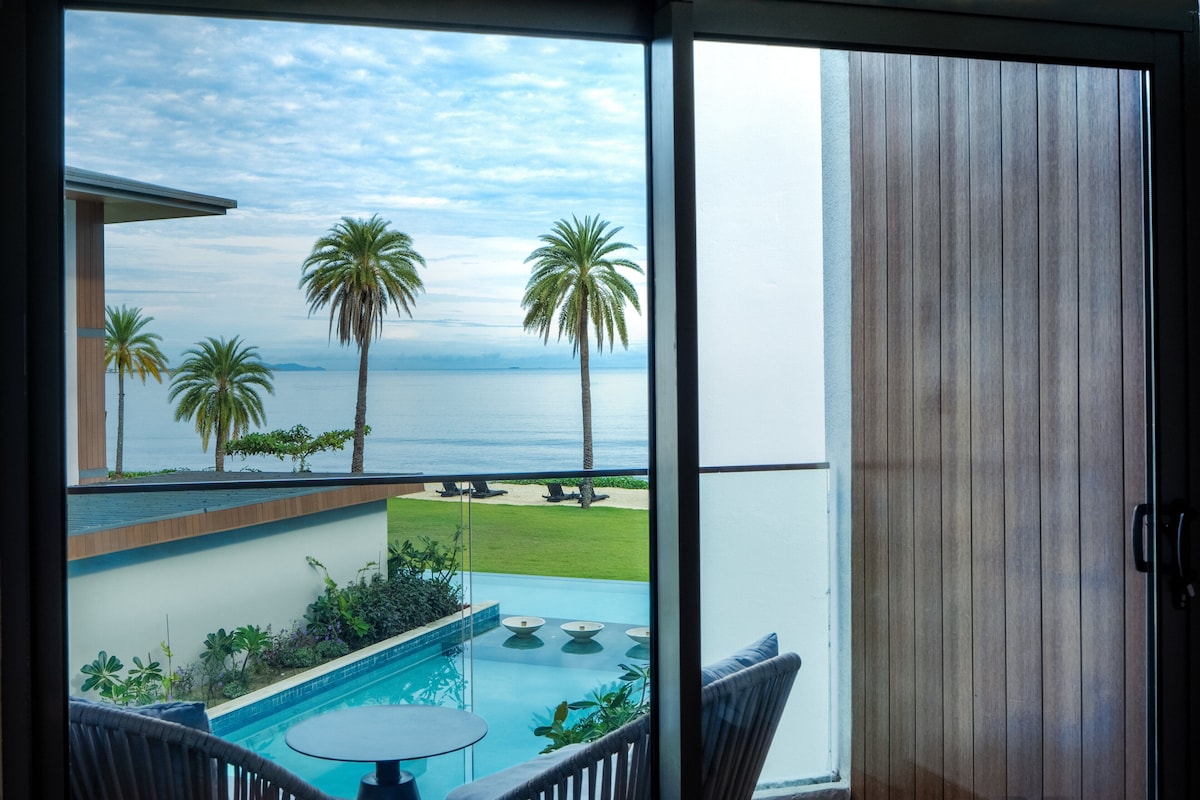 A balcony with a view of the sea, palm trees, and a swimming pool.