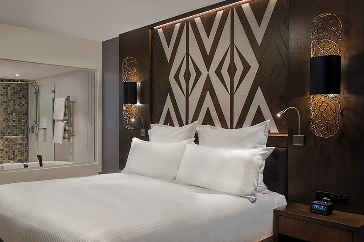 Una habitación de hotel moderna con una cama grande, un baño con ducha de vidrio y apliques decorativos en la pared.