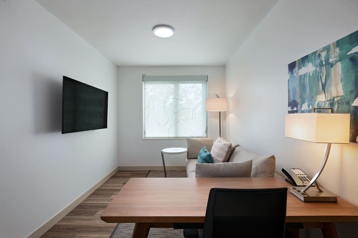 Une chambre d’hôtel moderne avec une table à manger en bois, un canapé, un téléviseur fixé au mur, une fenêtre donnant sur des arbres et un tableau accroché au mur.