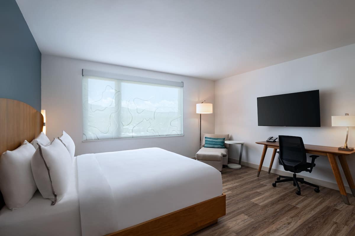 Une chambre d’hôtel avec un grand lit, un bureau avec une chaise, un téléviseur à écran plat et une fenêtre donnant sur le ciel.