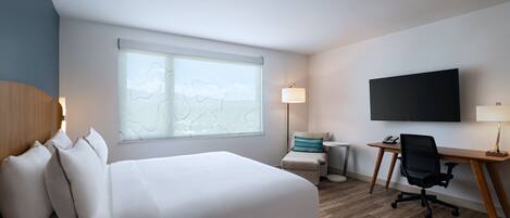 A hotel room with a large bed, a desk with a chair, a flat-screen TV, and a window with a view of the sky.