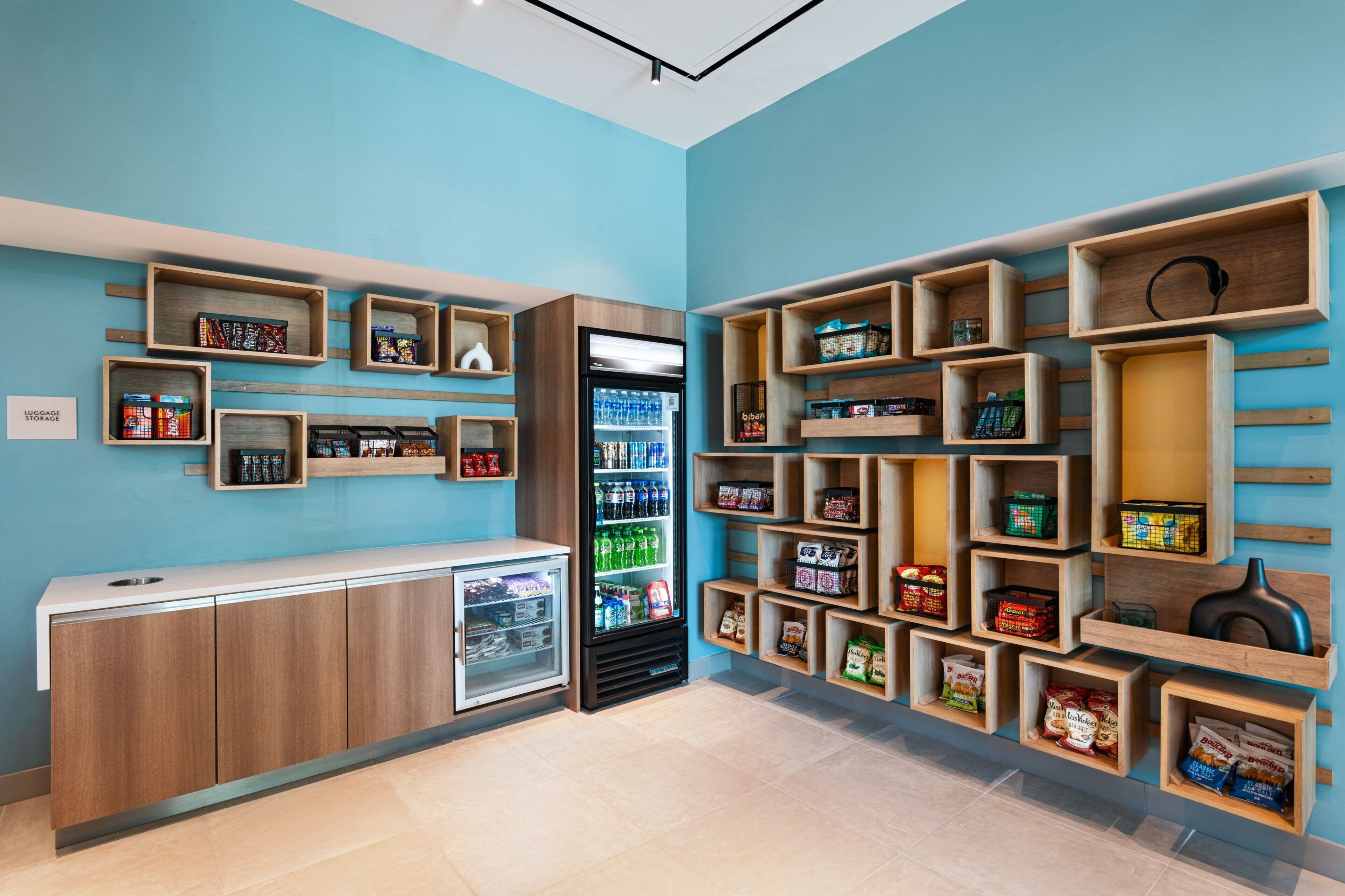 A hotel lobby with a blue wall, wooden shelves with snacks and beverages, a refrigerator, and a luggage storage area.