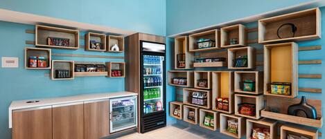 A hotel lobby with a blue wall, wooden shelves with snacks and beverages, a refrigerator, and a luggage storage area.