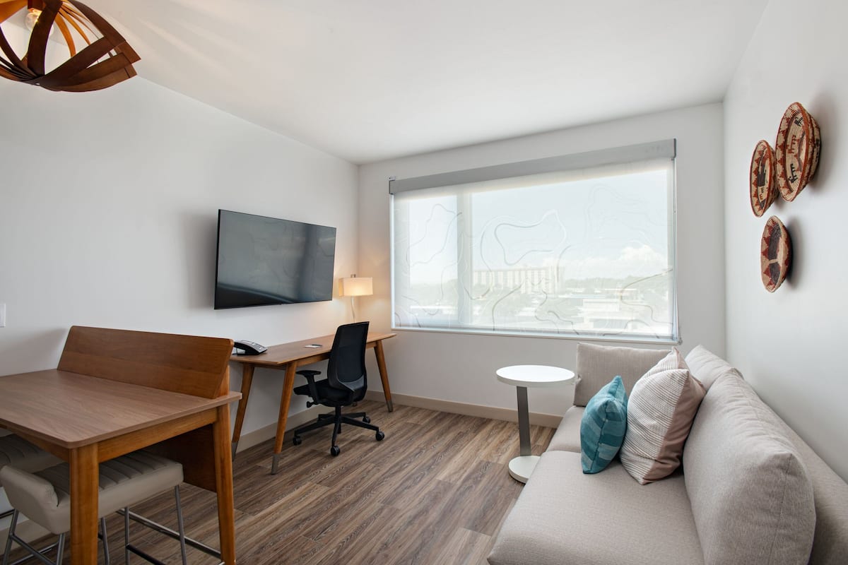 Une pièce moderne comprenant une table à manger en bois, un fauteuil de bureau noir, une petite table ronde blanche, un canapé beige agrémenté de coussins bleus et beiges, un téléviseur à écran plat fixé au mur et une grande fenêtre donnant sur un immeuble.