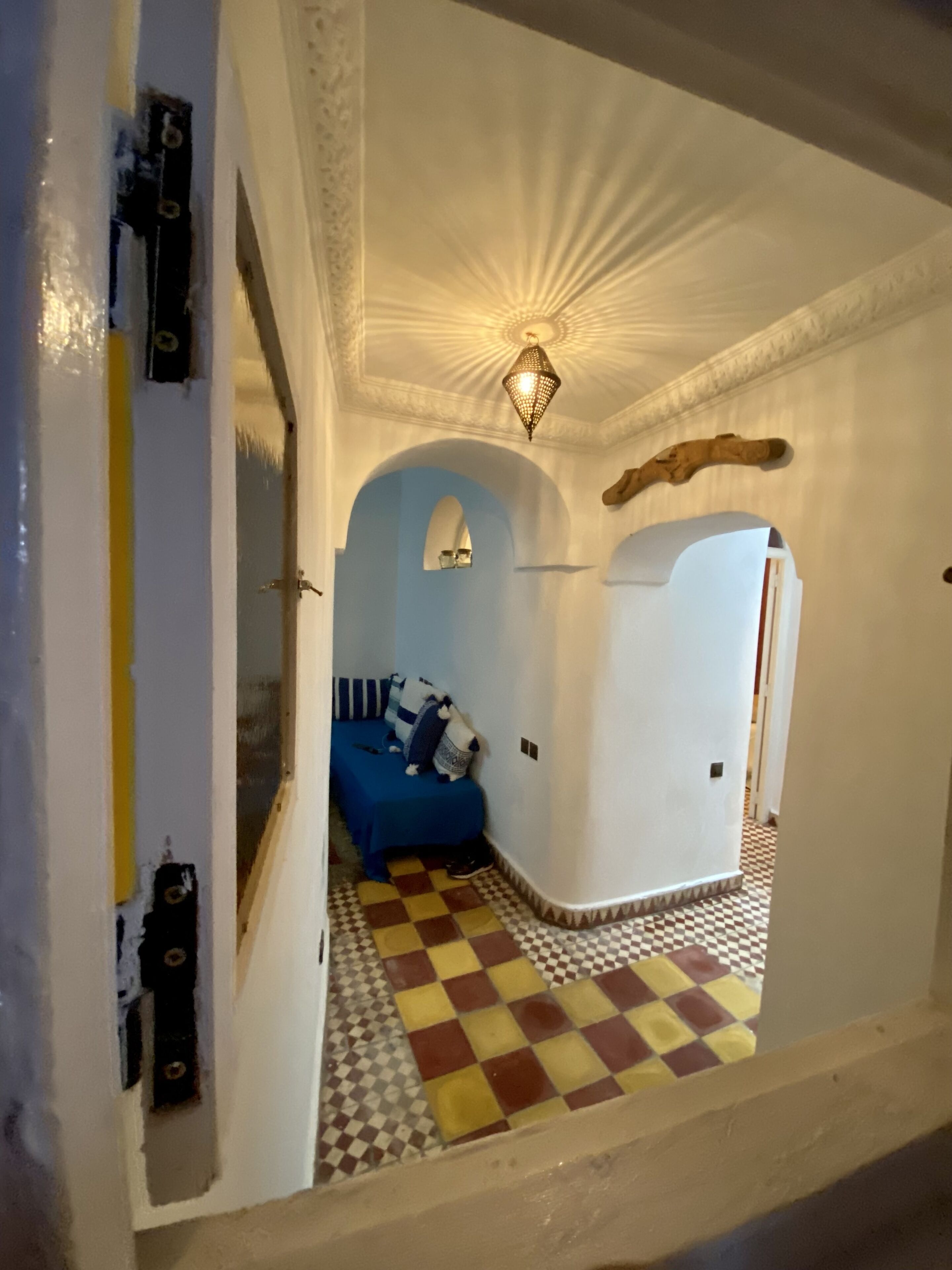 A narrow hallway with a checkered floor, a bed with a blue bedspread, and a decorative ceiling light.