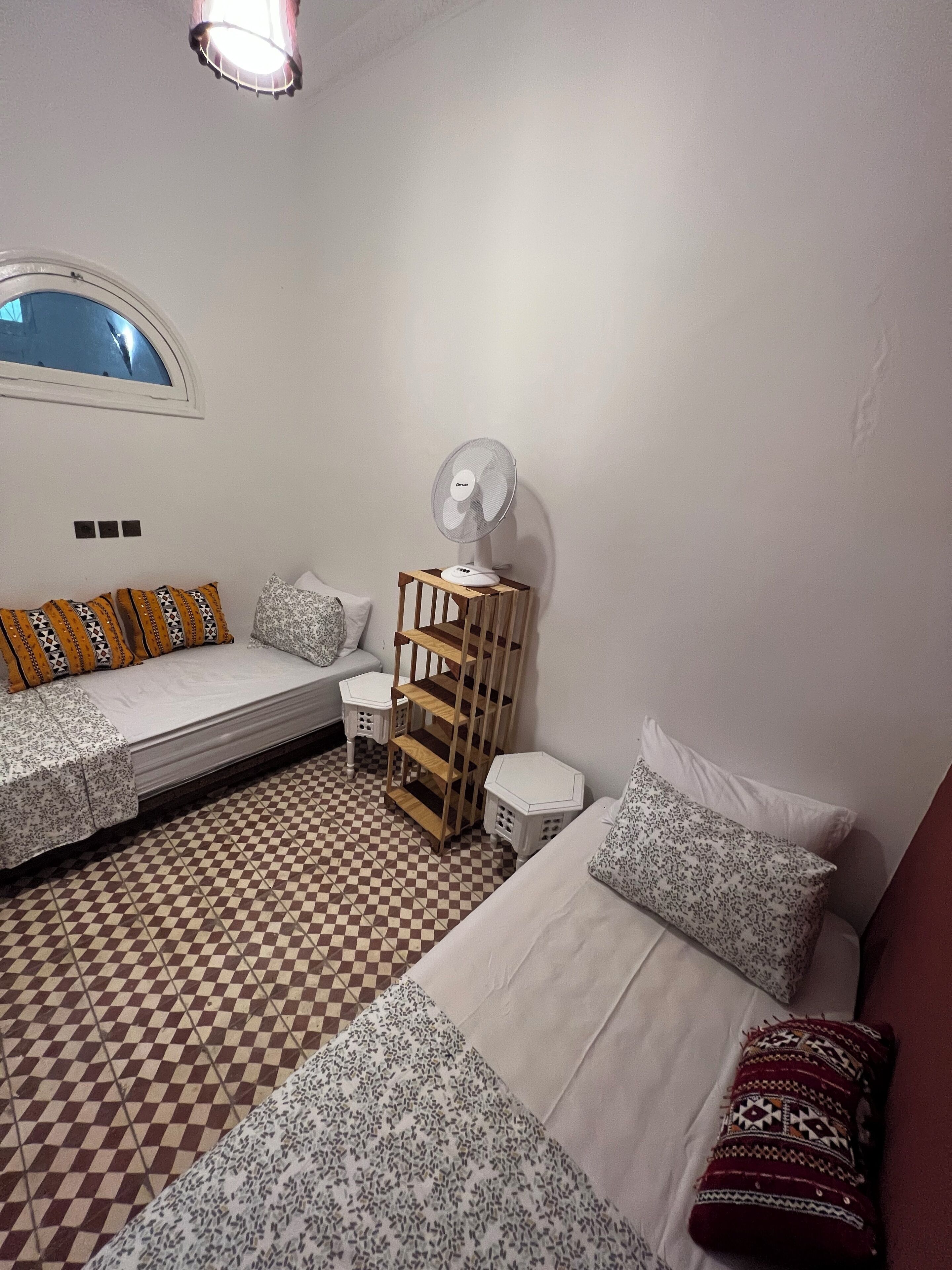 Una habitación con suelo a cuadros, dos camas con ropa de cama estampada, un ventilador de pie y una mesita.