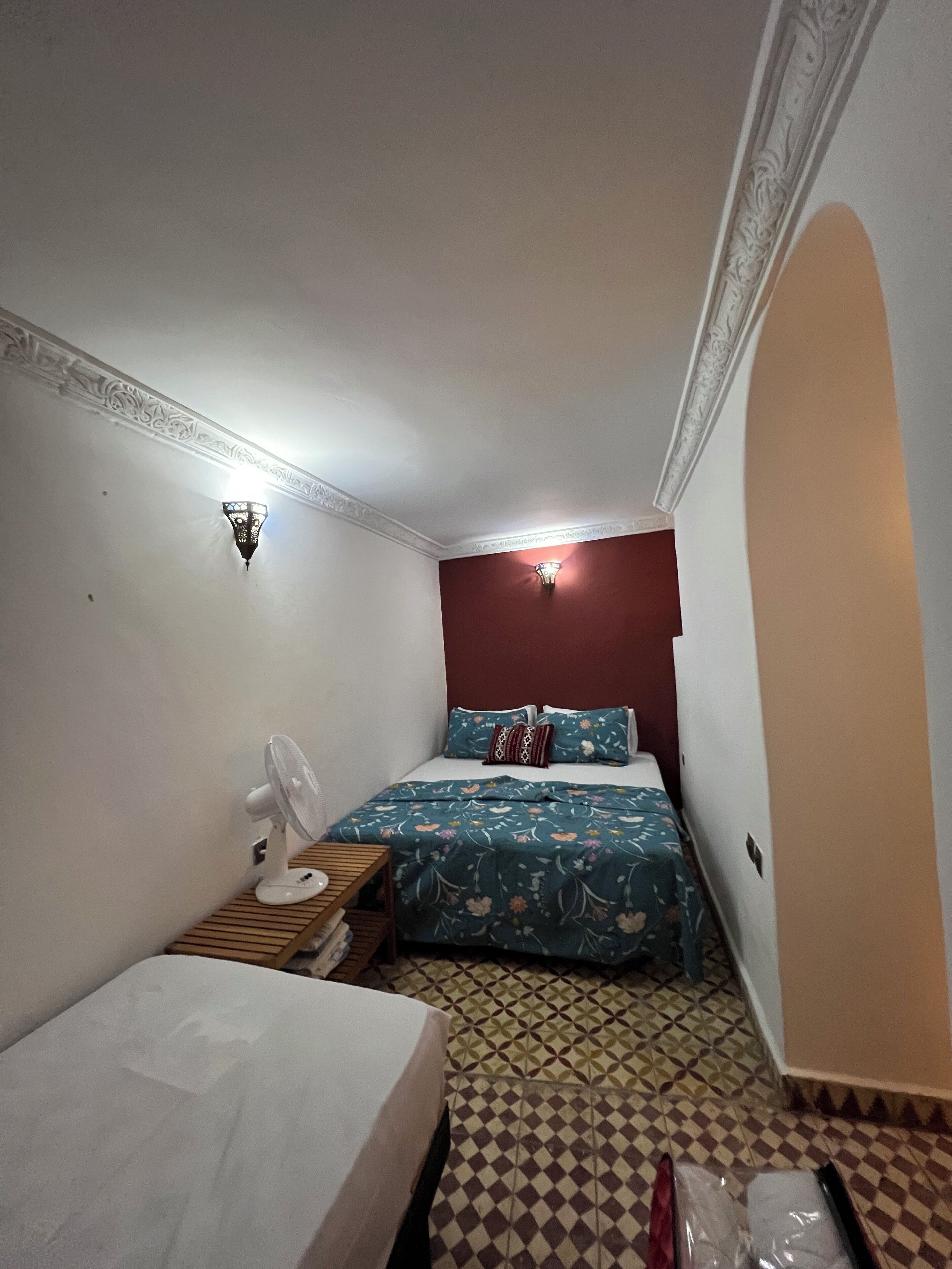 A small, compact room with a bed, a bedside table with a fan, a wall-mounted light, and patterned floor tiles.