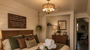 A bedroom with a large bed, a hanging plant, a bedside table with a lamp, and a wall-mounted clock.