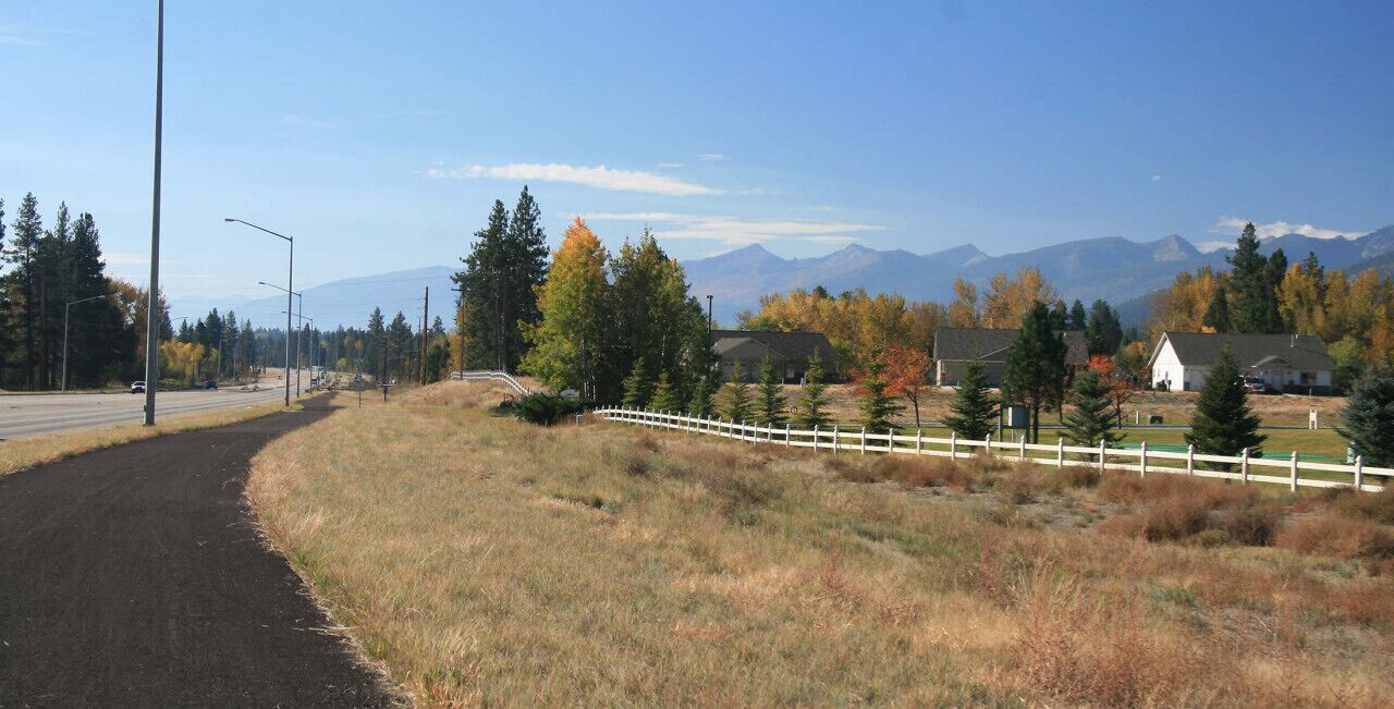 A rural road with a white fence, houses, and mountains in the background.