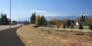 Une route de campagne bordée d’une clôture blanche, avec des maisons et des montagnes en arrière-plan.